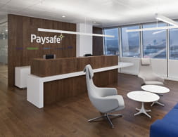 The reception area inside the Paysafe headquarters with a computer on top of a desk in front of a wood paneled wall with the company name Paysafe on it.