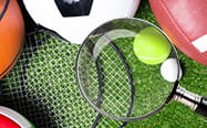 A pile of sports equipment including a tennis racket and ball, American football, football and basketball laying on a field with a magnifying glass.