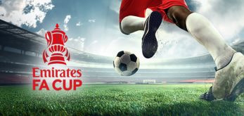 The image features a football player in motion on a grassy pitch, capturing the dynamic energy of a kick directed at a football. The Emirates FA Cup logo is prominently displayed on the left side of the image, set against the backdrop of a large, partially cloudy sky and an expansive stadium. This composition evokes the excitement and competitive spirit associated with the FA Cup tournament.