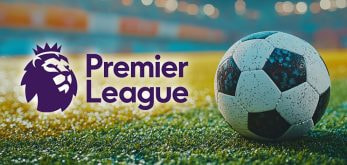 The image features a close-up view of a classic black and white soccer ball resting on a vibrant green grass pitch. To the ball's left is the Premier League logo, which includes a stylised purple lion's head accompanied by "Premier League" text in bold purple letters. The background displays a blurred stadium setting with bright lights, suggesting an atmosphere of excitement and anticipation for a football match.