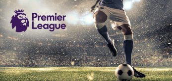 A dynamic action shot of a football player dribbling a ball on the field under stadium lights, with the Premier League logo prominently displayed, capturing the excitement and intensity of the game.