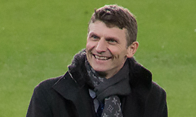 Tore André Flo smiling in a stadium, wearing a dark coat and patterned scarf, with a green football pitch in the background.