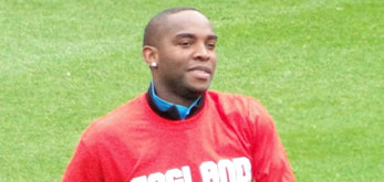 The image features Benni McCarthy wearing a red shirt with the word "ENGLAND" visible across the front. The individual appears to be on a grass field, possibly during a sports event. The background is out of focus, placing emphasis on the person in the foreground.