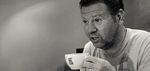 A black-and-white photo featuring a man sitting in a relaxed setting, holding a cup of coffee. He appears to be engaged in a conversation or deep in thought, creating a candid and natural moment. The background is blurred, keeping the focus on the man and his expression. The atmosphere of the photo is casual and intimate.